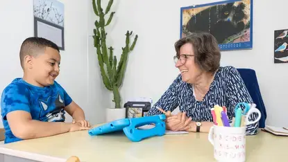 Leerkracht en leerling werken lachend samen aan een tafel met tablet.