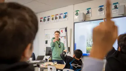 Leerkracht geeft les voor een digitaal schoolbord, leerling steekt zijn hand op.
