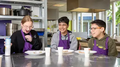 Drie leerlingen zitten lachend aan tafel in een keuken.