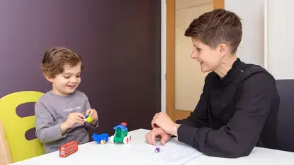 Een logopedist zit aan een tafel tegenover een peuter. Het kind speelt met speelgoedauto's en lacht. De logopedist kijkt toe en maakt aantekeningen.