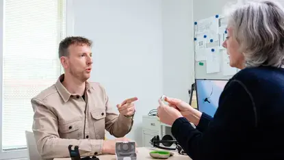 Een man overlegt met een audioloog. Op tafel liggen hoortoestellen en een doosje. De audioloog houdt een hoortoestel vast en legt iets uit.