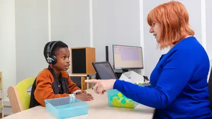 Een kind met grote koptelefoon doet een gehoortest. Tegenover hem zit een audioloog die de test begeleidt. Op tafel staan bakjes met gekleurde blokjes.