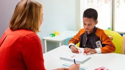 Een kind werkt met rode blokjes aan een taak. Tegenover hem zit een therapeut die aantekeningen maakt. Het kind is geconcentreerd bezig.