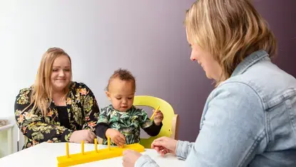 Een baby speelt met een geel speelgoedrek aan een tafel. Naast het kind zit de moeder. Tegenover hen zit een therapeut die het kind begeleidt.