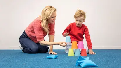 Een leerling bouwt een piramide van gekleurde bekers, met een begeleider ernaast.