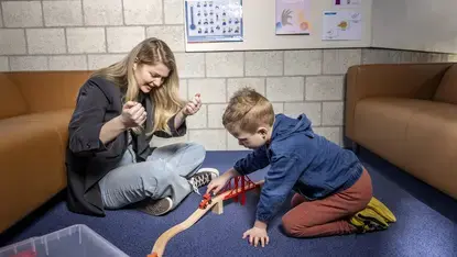 Een logopedist en een jongen spelen samen met een houten treinbaan en brug op de grond.