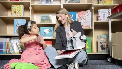 Een logopedist leest voor uit een prentenboek aan een meisje, tussen boekenkasten.