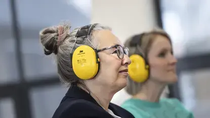 Een vrouw met een gele gehoorbeschermer kijkt naar voren, met een collega op de achtergrond.