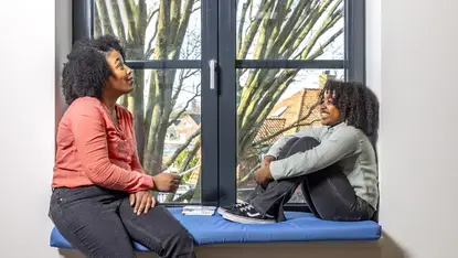 Een vrouw en kind zitten tegenover elkaar op een vensterbank met kaarten, in gesprek.