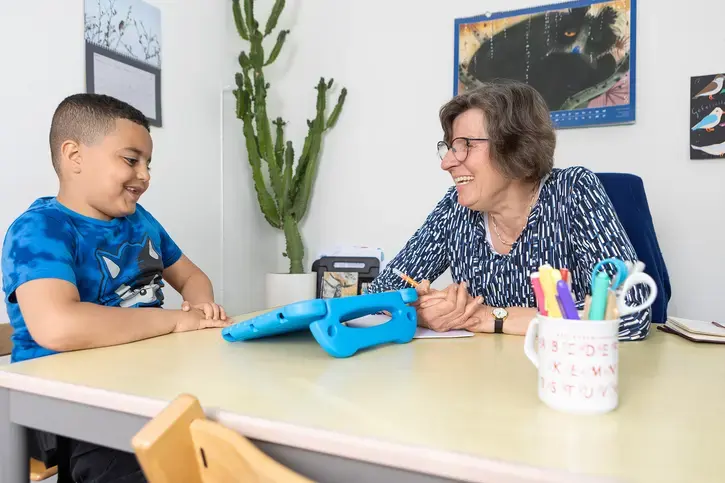 Leerkracht en leerling werken lachend samen aan een tafel met tablet.