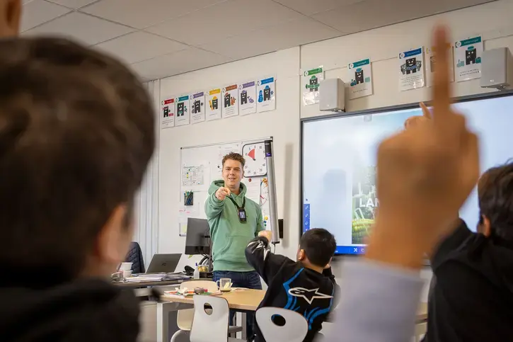 Leerkracht geeft les voor een digitaal schoolbord, leerling steekt zijn hand op.
