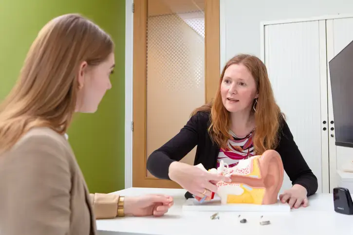 Een audioloog legt aan een jonge vrouw uit hoe het oor werkt. Ze wijst naar een groot anatomisch model van een oor. Op tafel liggen kleine hoortoestellen.