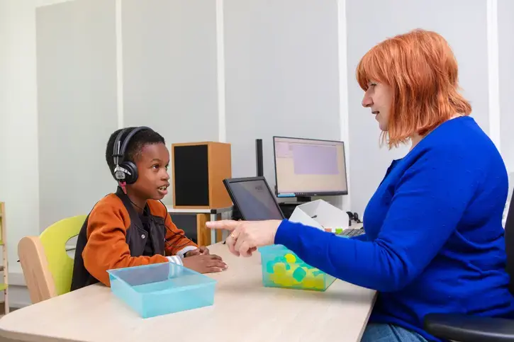 Een kind met grote koptelefoon doet een gehoortest. Tegenover hem zit een audioloog die de test begeleidt. Op tafel staan bakjes met gekleurde blokjes.