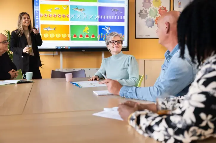 Een vrouw met bril lacht tijdens een training. Op de achtergrond staat een docent bij een digitaal scherm met kleurrijk lesmateriaal.