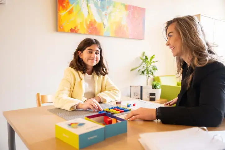Een meisje en een logopediste zitten tegenover elkaar aan een tafel met gekleurde blokken. Het meisje kijkt glimlachend naar de logopediste.