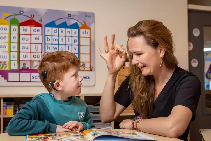 Een begeleider maakt een gebaar terwijl ze met een leerling met cochleair implantaat een prentenboek bekijkt.