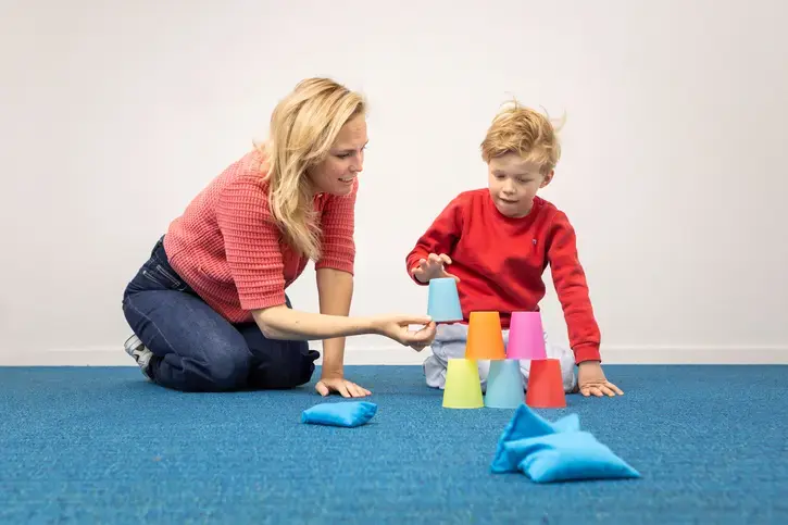 Een leerling bouwt een piramide van gekleurde bekers, met een begeleider ernaast.