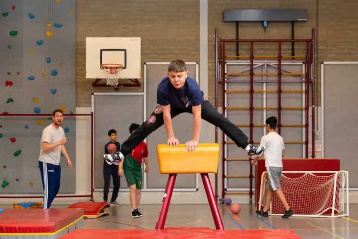 Een jongen maakt een spreidsprong over de pegasus in de gymzaal, terwijl een leraar en klasgenoten op de achtergrond toekijken.