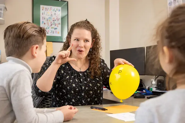 Een begeleider houdt een ballon met letters vast en legt iets uit aan twee leerlingen.