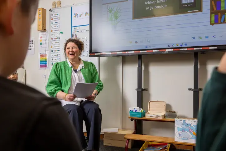 Een leerkracht leest lachend voor uit een boekje aan leerlingen in de klas.