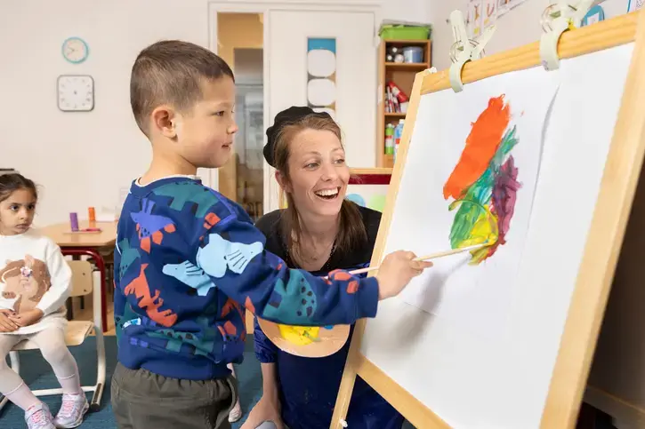Een leerling schildert op een ezel, met een lachende begeleider ernaast.