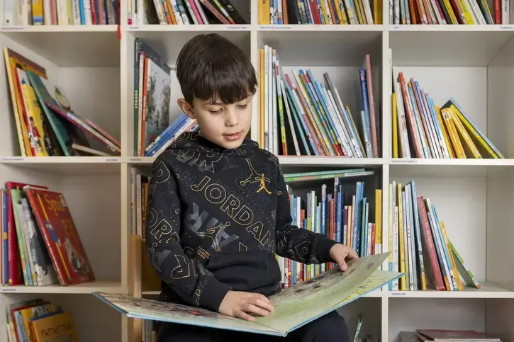 Een jongen leest een prentenboek voor een boekenkast met kinderboeken.