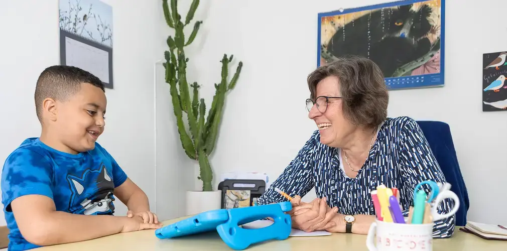 Leerkracht en leerling werken lachend samen aan een tafel met tablet.