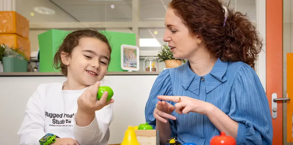 Meisje en leerkracht oefenen woordenschat met speelgoedfruit.