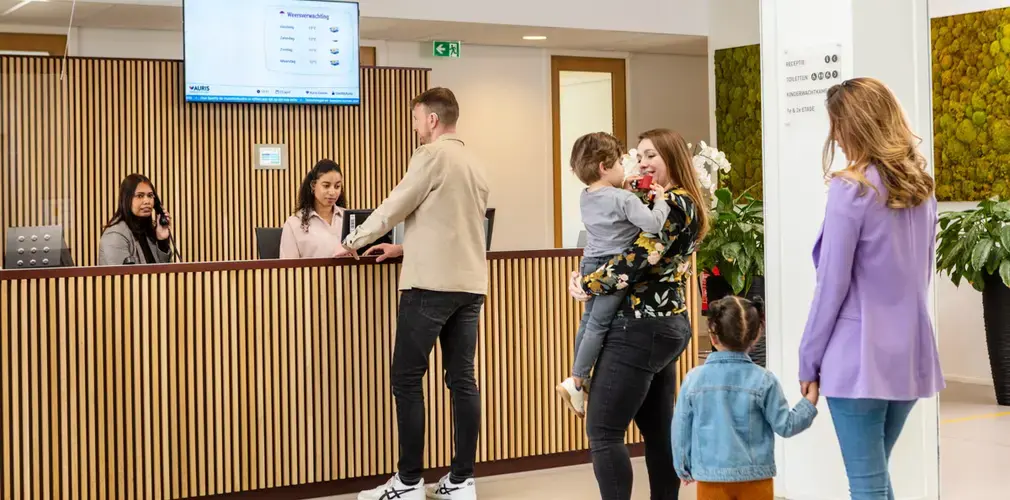 Twee families melden zich aan bij de receptiebalie van het Auris Audiologisch Centrum. Achter de balie staan twee medewerkers. Op een scherm is het Auris-intranet zichtbaar.