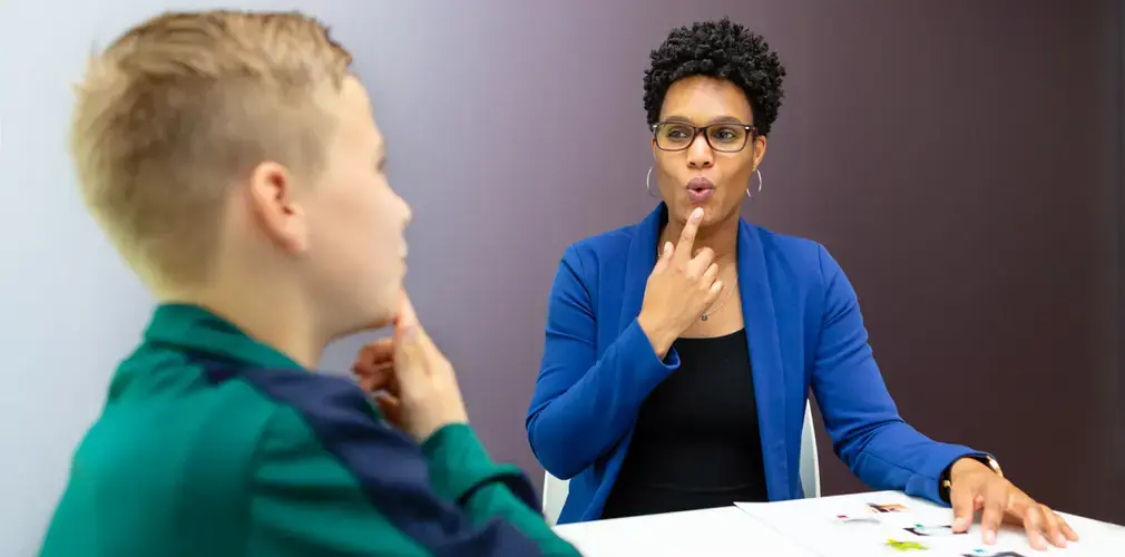 Een logopedist laat een jongen zien hoe hij zijn lippen moet bewegen. Ze wijst naar haar mond. Op tafel liggen kaartjes. De jongen kijkt haar na.