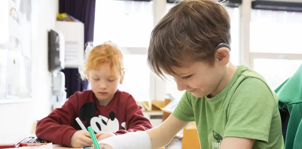Een leerling met hoortoestel schrijft aan zijn tafel in de klas.
