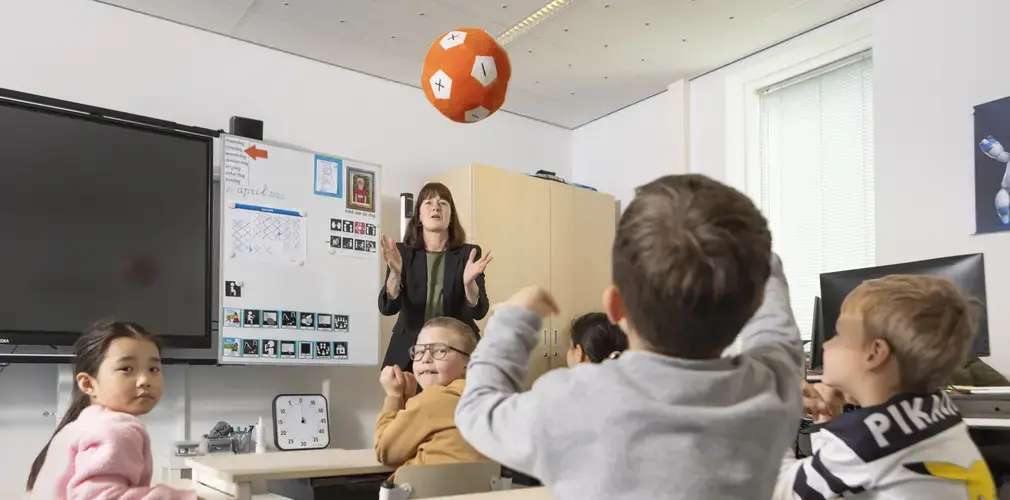 Leerkracht gooit een bal met rekentekens terwijl een groep leerlingen reageert in de klas.
