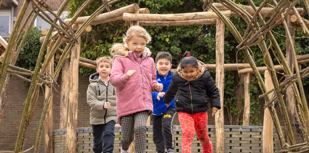 Vier leerlingen rennen lachend door een houten tunnel op het schoolplein.