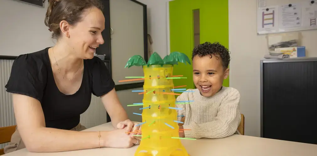 Een leerling doet een logopedie-oefening met een geel knutselwerk. De logopedist kijkt lachend toe.