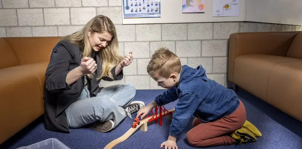 Een logopedist en een jongen spelen samen met een houten treinbaan en brug op de grond.