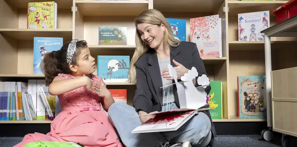Een logopedist leest voor uit een prentenboek aan een meisje, tussen boekenkasten.