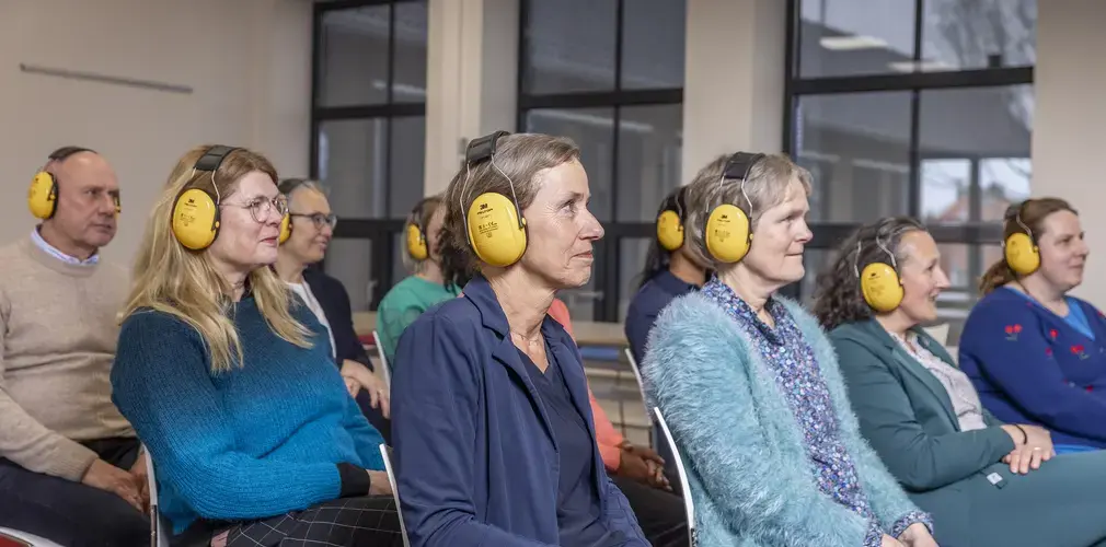 Een groep collega's met gele gehoorbeschermers luistert tijdens de cursus Ervaar Gehoorverlies..