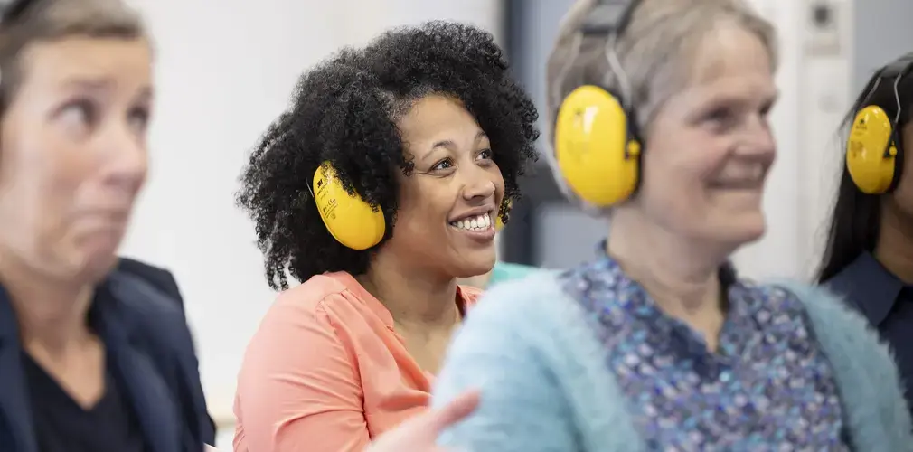 Een lachende vrouw met gele gehoorbeschermer, tussen collega's tijdens een workshop.