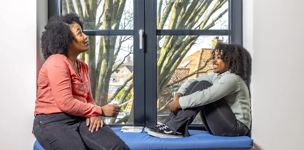 Een vrouw en kind zitten tegenover elkaar op een vensterbank met kaarten, in gesprek.
