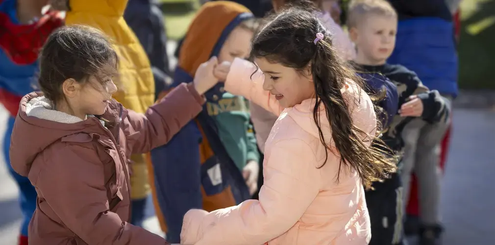 Twee meisjes stoten hun ellebogen tegen elkaar buiten, tussen andere kinderen.