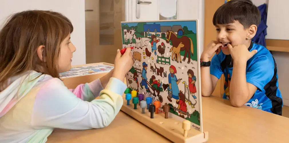 Twee kinderen spelen samen met een taalspel met een boerderijplaat en gekleurde pinnen aan tafel.