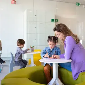 In een wachtruimte tekenen twee jonge kinderen aan ronde tafeltjes. Een begeleider helpt een meisje. Op de achtergrond zit een moeder toe te kijken.