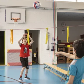 Twee leerlingen spelen samen een potje badminton in de gymzaal. Ze bewegen actief en lachen.