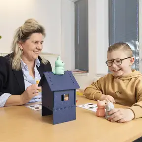 Een leerling speelt met een speelgoedhuisje en dierfiguren, met een begeleider aan tafel.