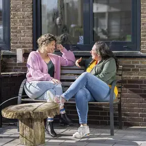 Twee vrouwen zitten buiten op een bankje in gesprek, in de zon.
