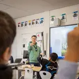 Leerkracht geeft les voor een digitaal schoolbord, leerling steekt zijn hand op.