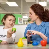 Meisje en leerkracht oefenen woordenschat met speelgoedfruit.
