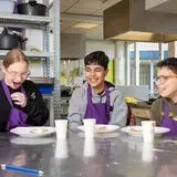 Drie leerlingen zitten lachend aan tafel in een keuken.