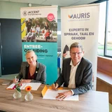 Twee mensen ondertekenen een overeenkomst aan een tafel, met banners van Auris en Accent op de achtergrond.