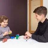 Een logopedist zit aan een tafel tegenover een peuter. Het kind speelt met speelgoedauto's en lacht. De logopedist kijkt toe en maakt aantekeningen.